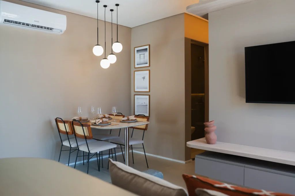 apartamento para financiar: Sala de estar e área de jantar em apartamento moderno para financiar, com mesa retangular, cadeiras, luminárias pendentes e painel de TV na parede, além de ar-condicionado e decoração minimalista.
